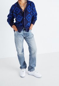 Marineblauer Cardigan mit blauen Diamantmustern, geknöpft, kombiniert mit hellblauen Jeans und weißen Sneakers, der einen lässigen Schnitt und eine weiche Textur aufweist.