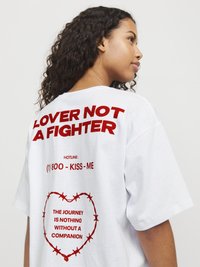 Hvit oversized t-skjorte med rødt trykt tekst på ryggen: "LOVER NOT A FIGHTER" og et hjerte omkranset av piggtråd.