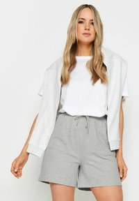 Shorts en coton gris clair avec cordon de serrage, associés à un t-shirt blanc à manches courtes et un sweat-shirt gris drapé sur les épaules.