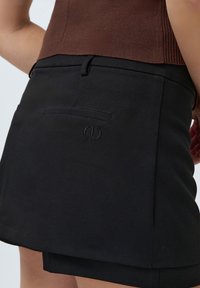 Fekete szoknya sima anyagból, hátsó zsebbel, vízszintes varrási részletekkel és diszkrét logóval. Az alja egyenes.