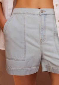 Shorts en denim bleu clair avec une taille haute, des poches avant et des ourlets retroussés. Le tissu semble doux avec des détails de couture visibles.