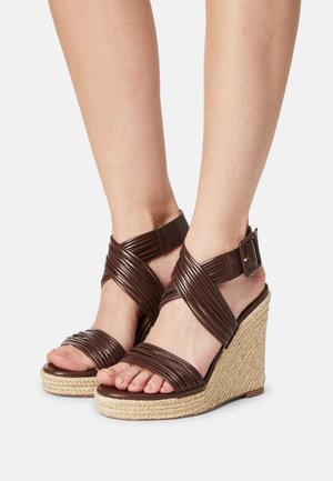 Sandales compensées marron avec des lanières croisées, fabriquées en matériau texturé. La semelle en jute tressé ajoute de la hauteur et un contraste au design.