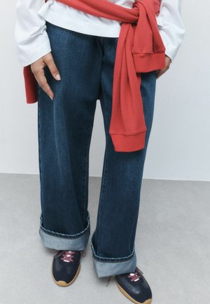 Person iført mørkeblå vidde bukser med opsmøgede manchetter, sorte sneakers med brune såler og pink snørebånd, en hvid top og en rød sweater bundet omkring livet.