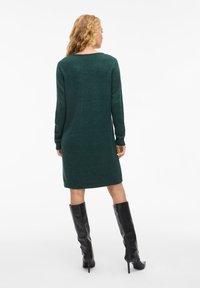 Abito maglione verde a maglia con maniche lunghe e scollo rotondo, caratterizzato da una vestibilità comoda e orlo a coste, abbinato a stivali neri fino al ginocchio.