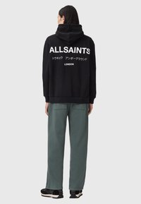 Czarna bluza z kapturem z białym napisem "ALLSAINTS" oraz japońskimi znakami z tyłu, połączona z luźnymi zielonymi spodniami i czarnymi sneakersami.