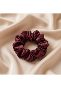 Chouchou en satin rouge foncé avec une petite étiquette "le silque" reposant sur un tissu satin lisse beige clair.