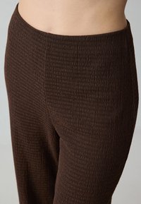 Pantalons en tissu côtelé marron avec une taille ajustée, présentant un motif de rayure verticale texturée. Le matériau semble doux et flexible.