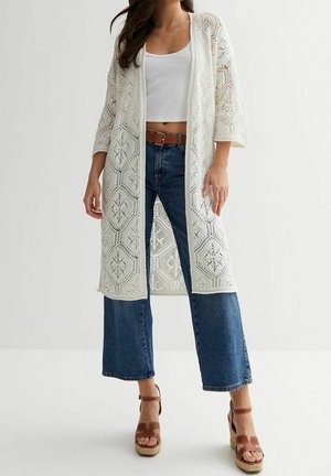 Frau trägt weiße gehäkelte lange Strickjacke über weißem Crop-Top, blaue Jeans mit weitem Bein, braunen Gürtel und braune Plateausandalen.