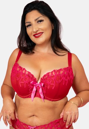 Smiling woman with long dark hair wearing red embroidered lingerie with pink ribbon detail, adjusting matching bottoms against plain background.