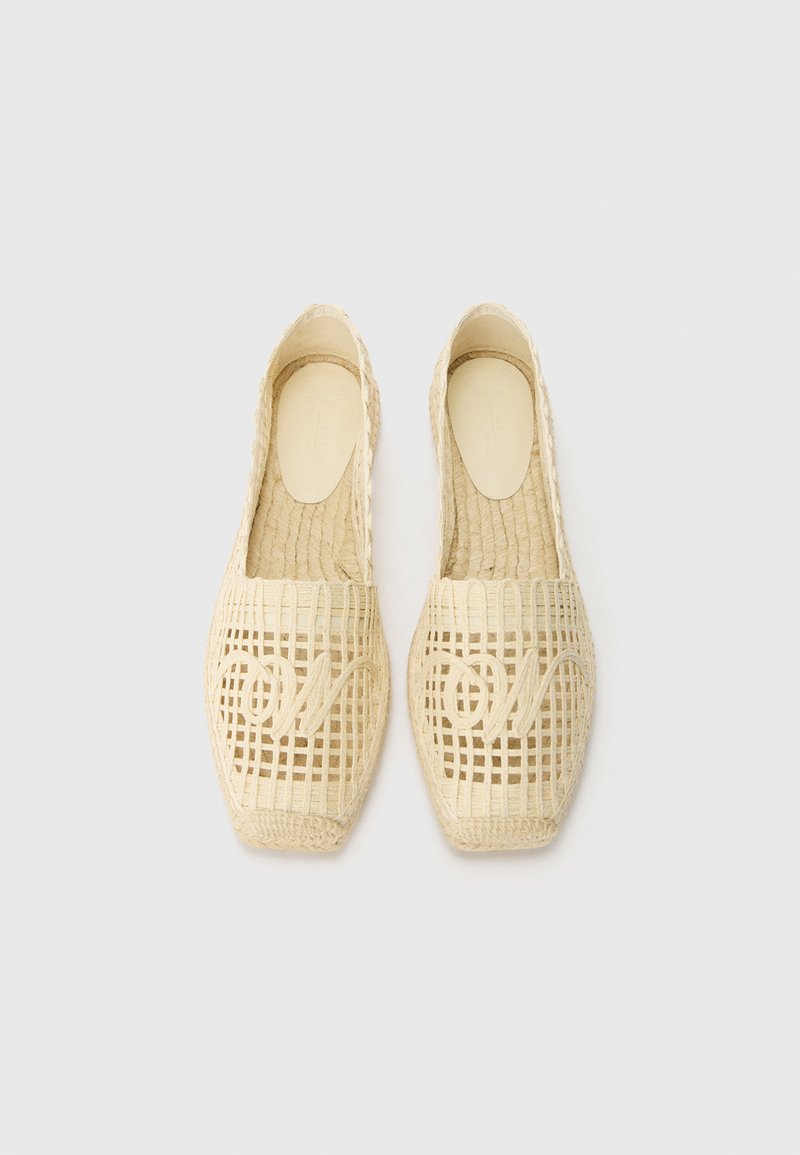 Woven, kremowe buty wsuwane z otwartym wzorem siatkowym oraz wyraźnym wzorem "o" na czubku, z płaską podeszwą i teksturowanym wykończeniem.