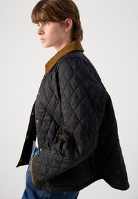 Jachetă neagră matlasată cu guler de catifea, dotată cu nasturi aurii și buzunare laterale, prezentată din profil pe un fundal neutru.