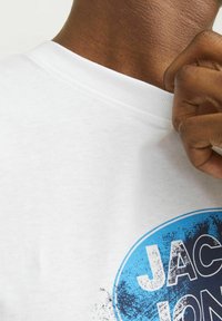 Hvid bomulds t-shirt med rund hals, der har et blåt grafisk design, som inkluderer tekst og et tekstureret mønster på forsiden.