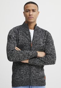 Suéter de punto negro y gris con cremallera, cuello alto, puños acanalados y un parche de la marca en el dobladillo. Presenta un patrón texturizado.