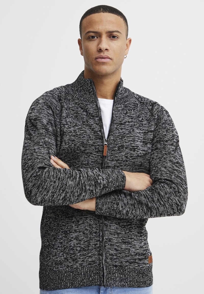 Suéter de punto negro y gris con cremallera, cuello alto, puños acanalados y un parche de la marca en el dobladillo. Presenta un patrón texturizado.