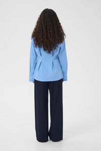 Φωτεινό γαλάζιο μακρυμάνικο πουκάμισο με εφαρμοστή σχεδίαση στην πλάτη, σε συνδυασμό με φαρδιά σκούρα μπλε παντελόνια, εμφανίζεται από την πίσω όψη.