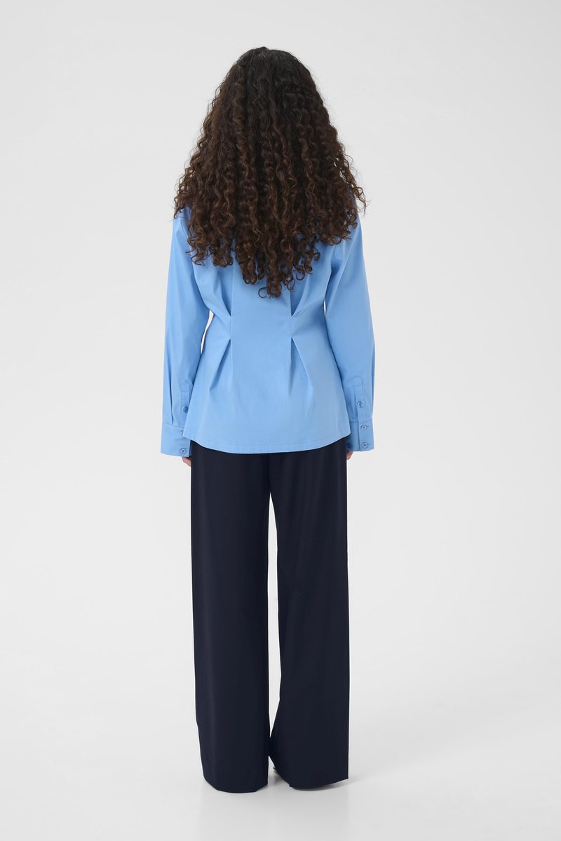 Chemise bleu clair à manches longues avec un dos cintré, associée à un pantalon ample de couleur bleu marine foncé, vue de dos.