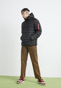 Giacca a vento nera con cappuccio, dotata di zip frontale e dettaglio rosso sulla manica sinistra. Pantaloni larghi marroni e sneakers bianche completano il look.