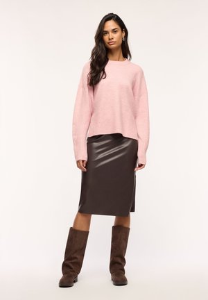 Donna che indossa un maglione rosa a maniche lunghe, una gonna in pelle marrone scuro al ginocchio con uno spacco laterale e stivali in camoscio marrone scuro, in piedi con sicurezza.