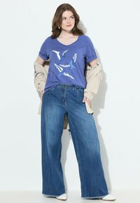 Camiseta morada con gráfico de pájaro, combinada con pantalones vaqueros de pierna ancha de mezclilla azul y un cárdigan beige. Zapatos blancos de punta acabada completan el conjunto.