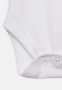 Bílý bavlněný bodysuit s krátkým rukávem, se zapínáním na patentky ve spodní části a hladkým, jemným povrchem. Nejsou viditelné žádné vzory ani detaily.