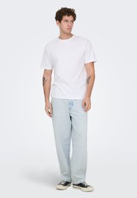 Λευκό βαμβακερό T-shirt με στρογγυλή λαιμόκοψη, σε συνδυασμό με ανοιχτό μπλε φαρδιά τζιν παντελόνια και μαύρα πάνινα αθλητικά παπούτσια. Το μοντέλο στέκεται με τα χέρια χαλαρά.