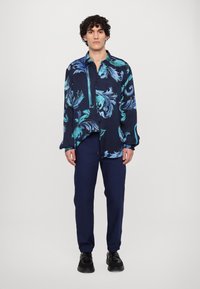 Marineblauw shirt met een gedurfde turquoise draaipatroon, lange mouwen, knoopsluiting, gecombineerd met marineblauwe broek en zwarte schoenen.