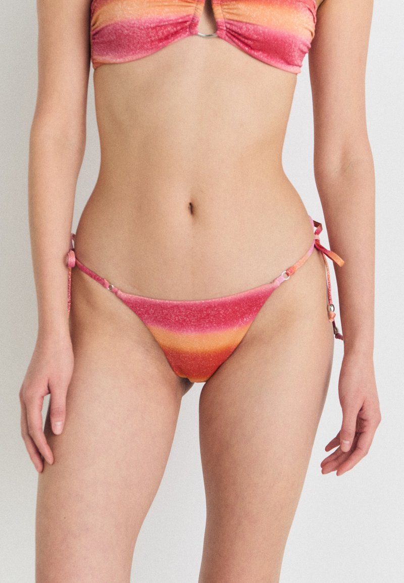 Bikini komplektas su gradientiniu raštu, derančiu rožinės ir oranžinės spalvos, su blizgiu tekstūra. Viršus turi centriniame žiede detalę; kelnaitės turi surišimo šonus.
