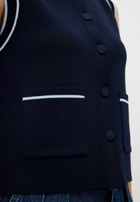 Gilet tricoté bleu marine avec un devant boutonné, deux poches avant avec bord blanc et un design court. Texture lisse, sans motifs supplémentaires.