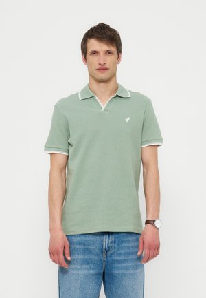 Άνδρας που φοράει ανοιχτοπράσινο πόλο με κοντά μανίκια και λευκή περιφέρεια, μπλε τζιν και ρολόι χειρός, στέκεται μπροστά σε απλό λευκό φόντο.