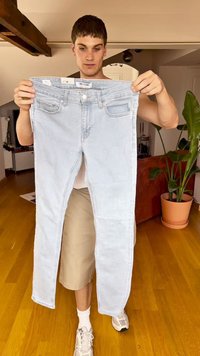 Calças de ganga azul claro seguradas verticalmente, com um corte slim, desbotado subtilmente, bolsos frontais e uma etiqueta da marca na cintura.