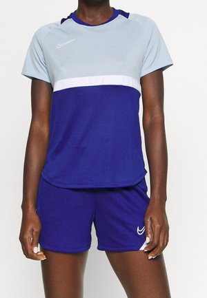 Kobieta ubrana w niebieską i jasnoszarą sportową koszulkę Nike oraz pasujące niebieskie spodenki, stoi z rękami swobodnie opuszczonymi wzdłuż ciała.