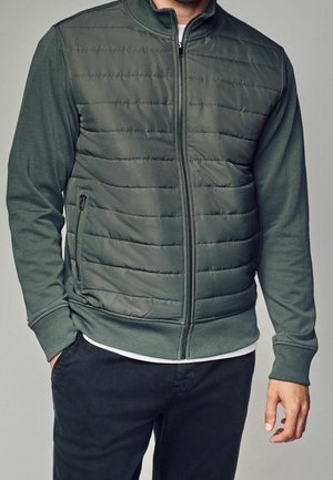 Homme portant une veste matelassée zippée vert foncé avec poignets et col côtelés, associée à un pantalon noir et un tee-shirt blanc.