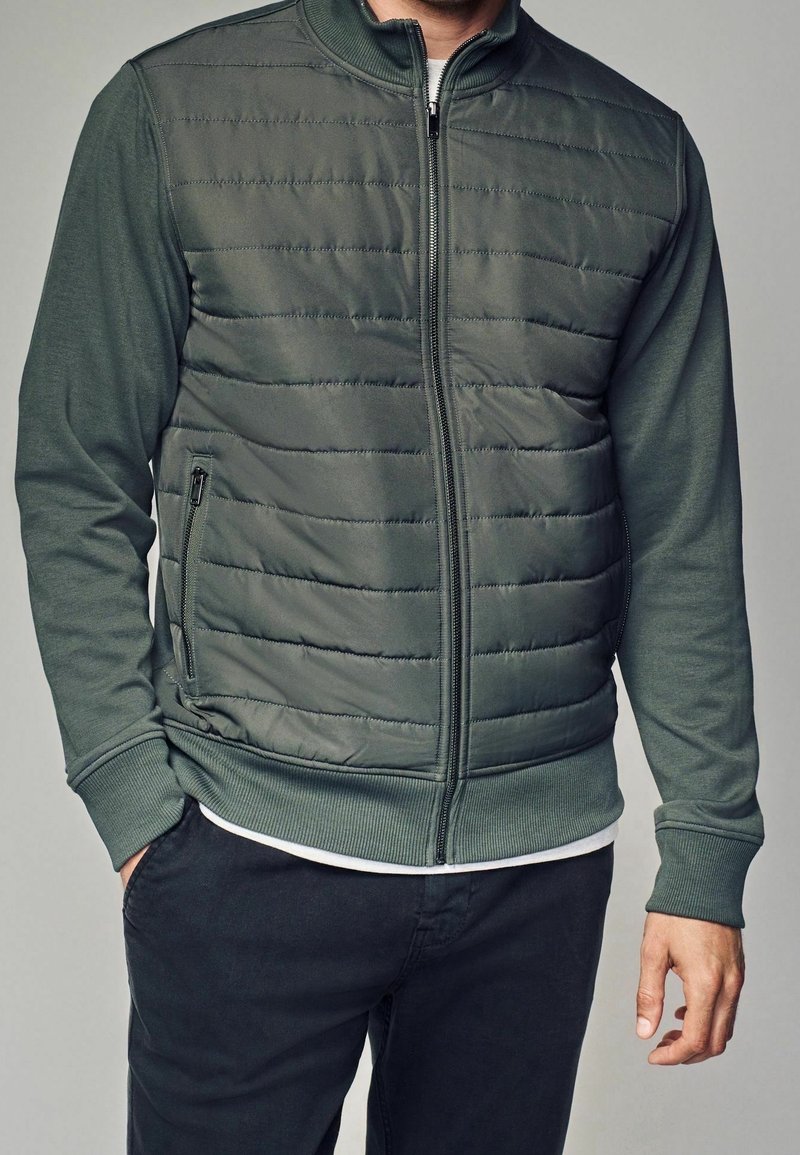 Homme portant une veste matelassée zippée vert foncé avec poignets et col côtelés, associée à un pantalon noir et un tee-shirt blanc.