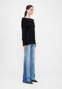 Schwarzer schulterfreier Pullover mit gerippter Textur, kombiniert mit hellblauen weiten Jeans, die kontrastierende Denim-Panele aufweisen, und schwarzen spitzen Schuhen.