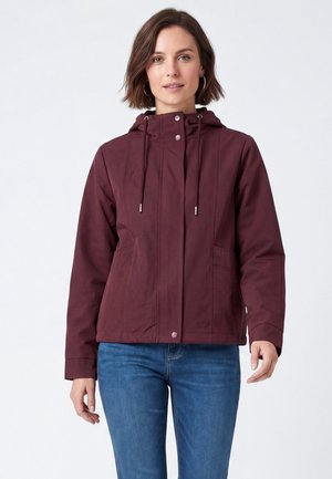 Donna che indossa una giacca con cappuccio color marsala con coulisse e bottoni a pressione, abbinata a jeans blu, in piedi davanti a uno sfondo bianco semplice.