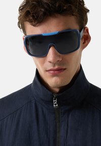 Lunettes de soleil avec une monture bleue, de grandes lentilles rectangulaires et un design distinctif sur la barre supérieure. Portées avec une veste noire à col montant.