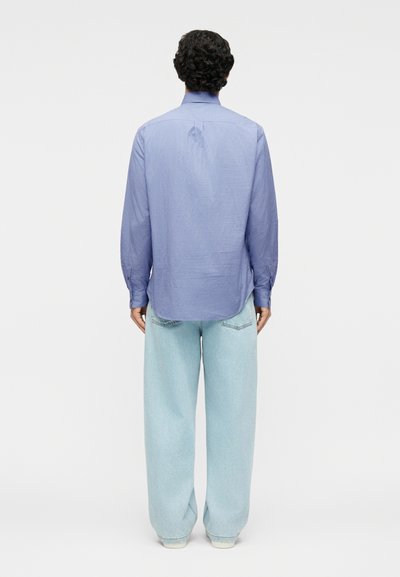 Άνδρας με σγουρά μαλλιά, που φοράει μπλε ριγέ μακρυμάνικο πουκάμισο και ανοιχτόχρωμα τζιν με φαρδύ πόδι, στέκεται με την πλάτη προς τον θεατή.