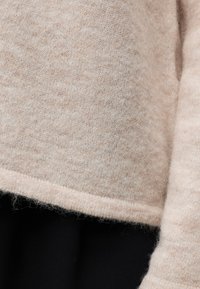 Manche et ourlet de pull en maille beige doux sur un pantalon noir, montrant la texture du tissu et les détails des coutures.