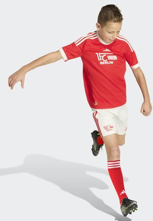 Niño con uniforme de fútbol del 1. FC Union Berlin en rojo, pateando el balón, con camiseta roja, pantalones cortos blancos, medias rojas y zapatillas negras, sobre un fondo liso.