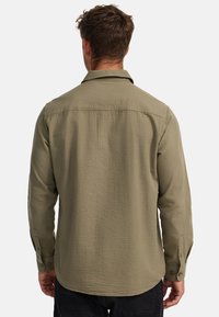 Olivgrünes, langärmliges Hemd aus einem strukturierten Stoff, mit Knopfmanschetten und klassischem Kragen. Sichtbare Nahtdetails an den Schultern.