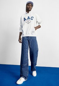 Pilkas medvilnės gobtuvėlis su "AAC" logotipu, derinamas su plačiomis mėlynomis džinsomis ir baltomis sportinėmis bateliais, fone esant mėlynai spalvai.