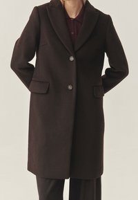 Manteau en laine brun foncé avec une coupe ajustée, col à revers crantés, deux boutons et poches latérales, présentant une texture lisse et une coupe droite.