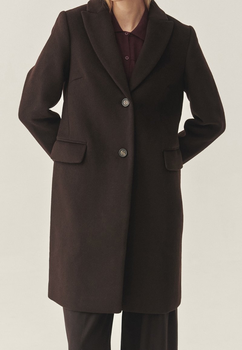 Manteau en laine brun foncé avec une coupe ajustée, col à revers crantés, deux boutons et poches latérales, présentant une texture lisse et une coupe droite.