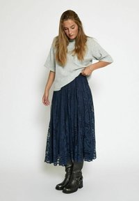Top grigio chiaro a maniche corte abbinato a una gonna maxi in pizzo blu navy con motivi intricati, indossato con stivaletti neri con tacco.