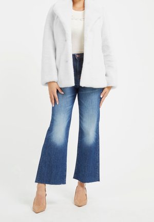 Nainen pukeutuneena valkoiseen pörröiseen takkiin, valkoiseen toppiin, korkeavyötäröisiin lyhennettyihin liehupöksyihin haalistuneilla polvilla ja beigen värisiin teräväkärkisiin korkokenkiin valkoisella taustalla.