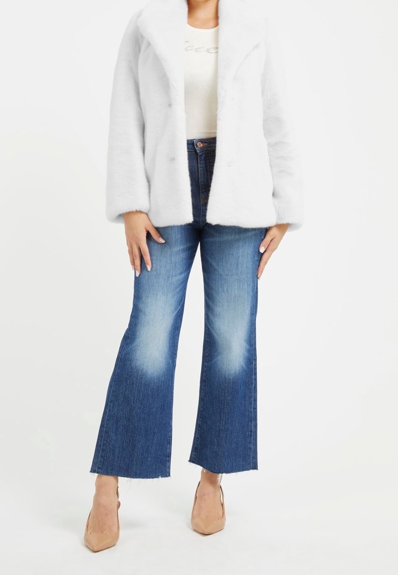 Femme portant une veste blanche duveteuse, un top blanc, un jean flare court taille haute avec genoux délavés et des talons beiges pointus sur fond blanc.