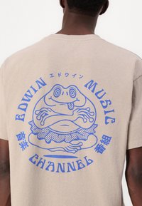 Beige t-skjorte laget av bomull med et blått grafikk av en frosk som har på seg et volangskjørt, omgitt av teksten "EDWIN MUSIC CHANNEL."