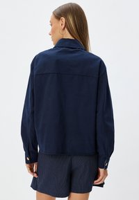 Camicia blu navy con colletto e maniche lunghe, dotata di polsini con bottone e orlo dritto. Il tessuto sembra liscio con texture minima.