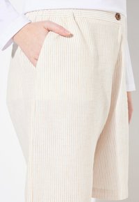 Béžové šortky s vertikálními světle hnědými pruhy, s boční kapsou a knoflíkovým detailem na pase. Hladká textura.