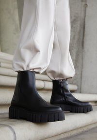 Zwarte leren enkellaarsjes met grove, getextureerde zolen en ritssluitingen aan de zijkant, gecombineerd met witte taps toelopende broek, getoond op stenen trappen.